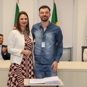 Um homem e uma mulher, de pé e um ao lado do outro, posam para foto atrás de uma mesa com um papel e uma caneta.