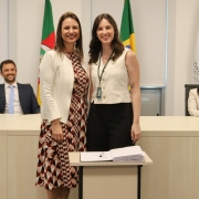 Duas mulheres, de pé e uma ao lado da outra, posam para foto atrás de uma mesa com um papel e uma caneta.