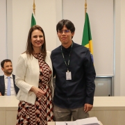 Um homem e uma mulher, de pé e um ao lado do outro, posam para foto atrás de uma mesa com um papel e uma caneta.