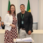 Um homem e uma mulher, de pé e um ao lado do outro, posam para foto atrás de uma mesa com um papel e uma caneta.