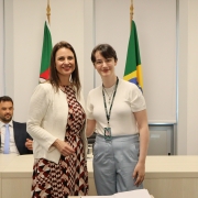 Duas mulheres, de pé e uma ao lado da outra, posam para foto atrás de uma mesa com um papel e uma caneta.