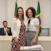 Duas mulheres, de pé e uma ao lado da outra, posam para foto atrás de uma mesa com um papel e uma caneta.