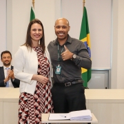 Um homem e uma mulher, de pé e um ao lado do outro, posam para foto atrás de uma mesa com um papel e uma caneta.