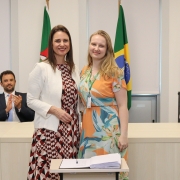 Duas mulheres, de pé e uma ao lado da outra, posam para foto atrás de uma mesa com um papel e uma caneta.