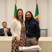 Um homem e uma mulher, de pé e um ao lado do outro, posam para foto atrás de uma mesa com um papel e uma caneta.