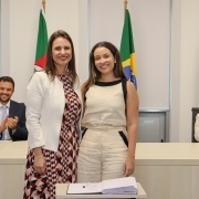 Duas mulheres, de pé e uma ao lado da outra, posam para foto atrás de uma mesa com um papel e uma caneta.