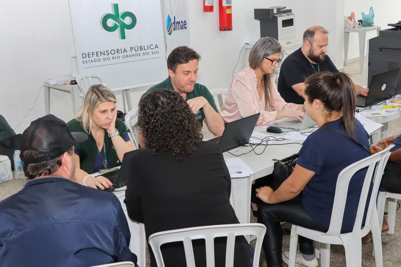 1 homem e 2 mulheres sentados a mesas e sendo atendidos por outras 2 mulheres e 2 homens do outro lado das mesas. Ao fundo, um cartaz com o símbolo da Defensoria Pública do Rio Grande do Sul e o nome da instituição.