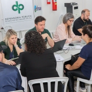 1 homem e 2 mulheres sentados a mesas e sendo atendidos por outras 2 mulheres e 2 homens do outro lado das mesas. Ao fundo, um cartaz com o símbolo da Defensoria Pública do Rio Grande do Sul e o nome da instituição.