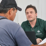 De costas para a foto, um homem sentado a uma cadeira olha para um papel que está sendo mostrado por outro homem, este de frente para a imagem e sentado à mesma mesa que ele.