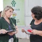 Duas mulheres, de pé e de lado para a foto, conversam. Uma delas está com uma prancheta e a outra segura um papel e seu celular.