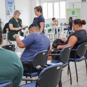 Pessoas de costas para a foto sentadas em cadeiras em uma sala fechada. Ao fundo, uma mulher é atendida por uma mulher com uma prancheta.