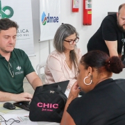 De costas para a foto, uma mulher está sentada em frente a uma mesa, à qual também estão sentados 2 homens e uma mulher que mexem em computadores. 