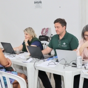 De costas para a foto, uma mulher está sentada em frente a uma mesa, à qual também estão sentados 2 mulheres e um homem que mexem em computadores. 