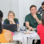 De costas para a imagem, 2 mulheres estão sentadas a uma mesa, à qual também estão sentados 2 homens e uma mulher que, do outro lado da mesa, mexem em computadores.