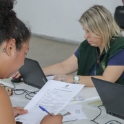 De costas para a foto, uma mulher olha para um papel e uma caneta que está segurando. Ao fundo, sentada a uma mesa, uma outra mulher mexe em um computador.