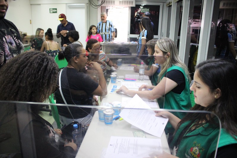 Pessoas sendo atendidas em mesas de atendimento por funcionários de colete verde.