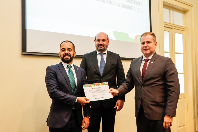 Na foto, há três homens em pé,  o defensor público-geral federal, Leonardo Cardoso de Magalhães; o subdefensor público-geral federal, Marcos Paderes; e o defensor público-geral do Estado, Nilton Leonel Arnecke Maria, segurando a homenagem recebida pela DPE/RS