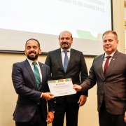 Na foto, há três homens em pé,  o defensor público-geral federal, Leonardo Cardoso de Magalhães; o subdefensor público-geral federal, Marcos Paderes; e o defensor público-geral do Estado, Nilton Leonel Arnecke Maria, segurando a homenagem recebida pela DPE/RS