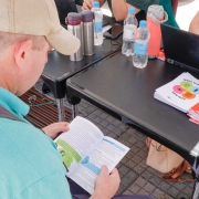 Homem sentado em uma cadeira, de costas para a foto, manuseia um caderno pequeno de papel e o lê. Em sua frente, uma pessoa manuseia um computador em uma mesa com garrafas de água e outros cadernos.