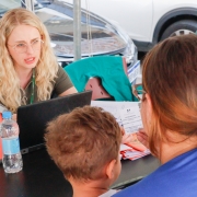 Em primeiro plano e de costas para a foto, uma mulher com uma criança de colo escuta uma outra mulher que, de frente para ela e para a foto, está falando enquanto manuseia um computador em uma mesa.