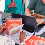 Uma mulher, de costas para a foto, manuseia livros pequenos de papel. Em um deles, pode-se ler o título "anticapacitismo para crianças". À frente dela, uma pessoa com camisa da Defensoria Pública do Rio Grande do Sul está sentada e manuseia um computador em uma mesa.