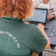 De costas para a foto, uma mulher com camisa da Defensoria Pública do RS manuseia um computador em uma mesa.