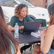 Uma mulher, de costas para a foto e sentada, fala com outra mulher que, também sentada, está de frente para ela e para a foto.