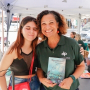 Duas mulheres, uma ao lado da outra, posam para a foto. Elas estão embaixo de tendas em um ambiente externo, urbano. A mulher da direita segura um livro em suas mãos.