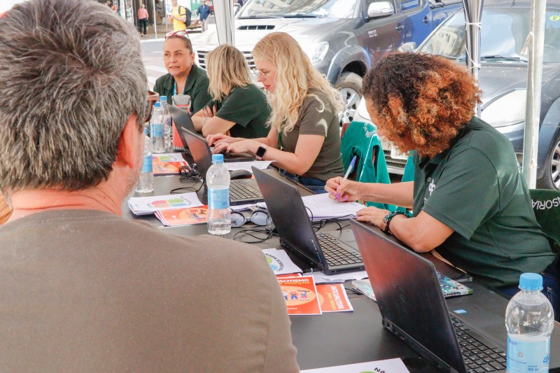 De costas para a foto, um homem olha para mulheres que estão em mesas de atendimento em uma área externa, mexendo em computadores e conversando.