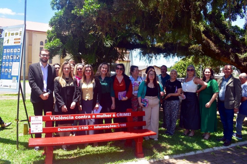 Grupo de pessoas de pé em uma praça posando para a foto. à frente delas, está no chão um banco vermelho com o escrito "violência contra a mulher é crime".