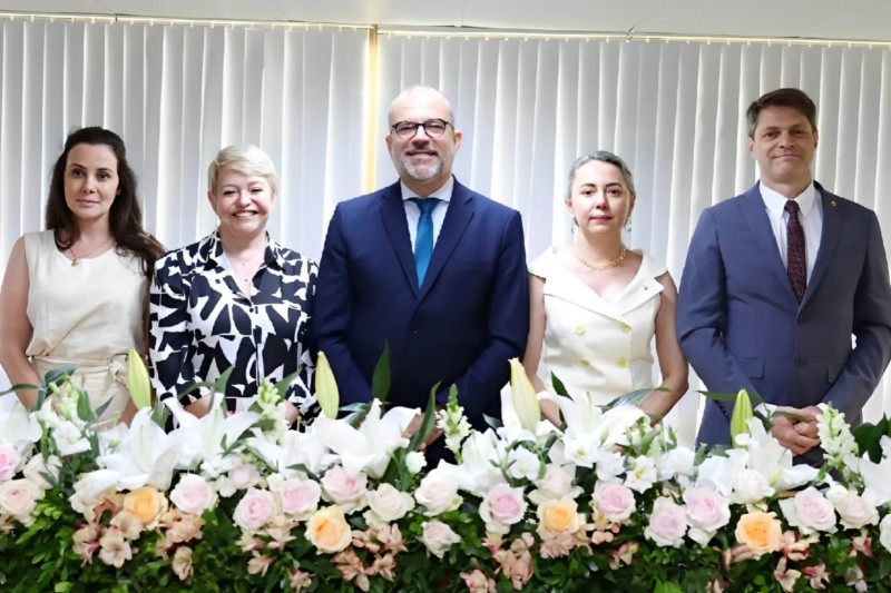 3 mulheres e 2 homens, todos de pé e um do lado do outro, estão posando para a foto dentro de uma sala. Em frente a eles, há várias flores em uma bancada, e atrás é possível ver uma cortina.