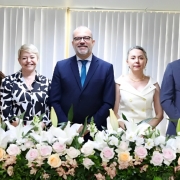 3 mulheres e 2 homens, todos de pé e um do lado do outro, estão posando para a foto dentro de uma sala. Em frente a eles, há várias flores em uma bancada, e atrás é possível ver uma cortina.