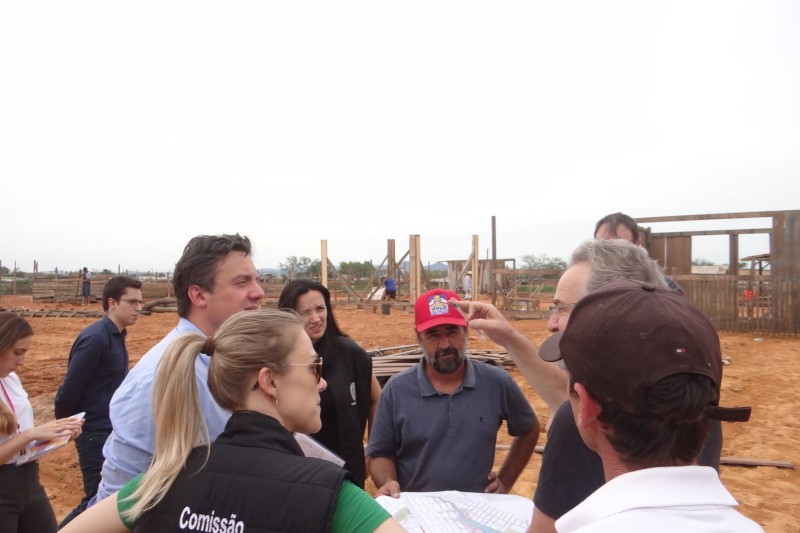 Grupo de 8 pessoas em um terreno de terra ao ar livre com algumas estruturas de madeira espaçadas.