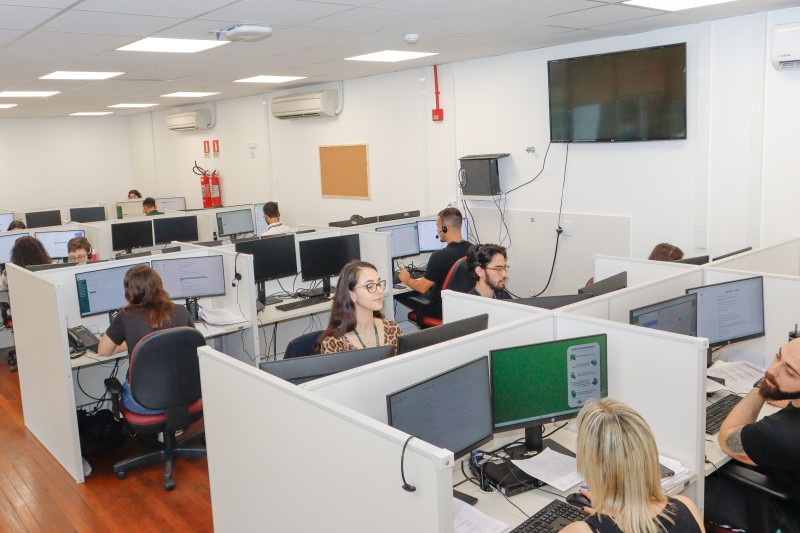 Sala de escritório com várias pessoas em cabines mexendo em computadores.