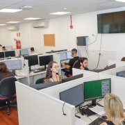 Sala de escritório com várias pessoas em cabines mexendo em computadores.