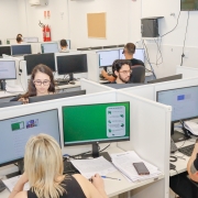 Sala de escritório com várias pessoas sentadas em cabines mexendo em computadores.