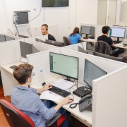 Várias pessoas sentadas a mesas e mexendo em computadores em cabines de uma sala de escritório.