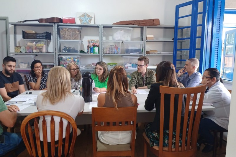 11 pessoas sentadas a uma mesa em uma sala rústica, conversando entre si com vários papéis e canetas na mesa.