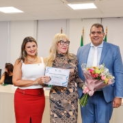 2 mulheres e um homem, de pé, estão de pé para a foto em uma sala branca fechada. Atrás deles, há uma mesa de palestra com pessoas. A mulher que está no meio dos outros 2 segura um certificado e um buquê de flores.