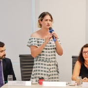 3 pessoas em uma mesa de palestra em uma sala branca. A pessoa que está no meio é uma mulher e está de pé, enquanto o homem e a mulher que estão ao seu lado estão sentados. A que está de pé, no meio, fala ao microfone.