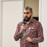 Um homem, de pé em uma sala branca e com uma bandeira LGBTQIAPN+ no peito, fala ao microfone em uma sala.