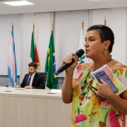 Uma mulher, de pé, fala ao microfone. Ao fundo, em uma mesa de palestra, estão sentadas 2 pessoas, havendo atrás delas bandeiras do Brasil, do Rio Grande do Sul e a bandeira trans