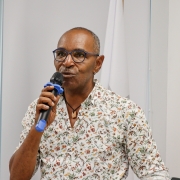 Um homem fala ao microfone de frente para a foto. Atrás dele, é possível ver uma bandeira do Brasil e outra bandeira LGBTQIAPN+.