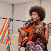 Uma mulher, sentada em um banco, toca um violão enquanto canta. À frente dela, uma estrutura segura um microfone e nela está apoiada uma bandeira LGBT. Ao fundo, na sala, é possível ver uma bandeira trans.