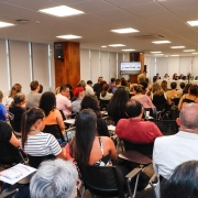 Um público de várias pessoas sentadas em cadeiras em uma sala branca.