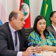 Um homem de terno, sentado em uma cadeira a uma mesa de palestra, fala ao microfone. Uma mulher e um homem ao seu lado aparecem desfocados na foto, também sentados à mesma mesa. Ao fundo, é possível ver bandeiras do Brasil, do Rio Grande do Sul e a bandeira trans.