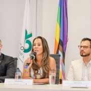 Uma mulher, sentada em uma cadeira a uma mesa de palestra, fala ao microfone. De cada um de seus lados, sentados à mesma mesa, há 2 homens de terno que olham para a mesma direção que ela. Ao fundo, é possível ver bandeiras do Brasil, do Rio Grande do Sul e a LGBTQIAPN+.