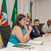 Uma mulher, em primeiro plano, fala ao microfone enquanto sentada a uma mesa de palestra, aparecendo de lado na foto. 2 homens, também sentados à mesma mesa e em segundo plano, olham para ela. Atrás deles, é possível ver bandeiras do Brasil, do Rio Grande do Sul e as bandeira trans e LGBTQIAPN+.