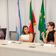 Uma mulher fala com um microfone na mão enquanto sentada a uma mesa de palestra, Outra mulher e também um homem, sentados à mesma mesa, olham para ela enquanto ela fala. Atrás deles, é possível ver bandeiras do Brasil, do Rio Grande do Sul e a bandeira trans