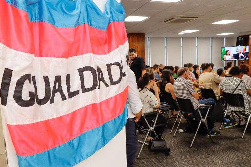 Em primeiro plano, uma bandeira transexual com o escrito "igualdade" no centro em uma porta branca. Em segundo plano, uma sala com várias pessoas sentadas em cadeiras e olhando para uma mesma direção.